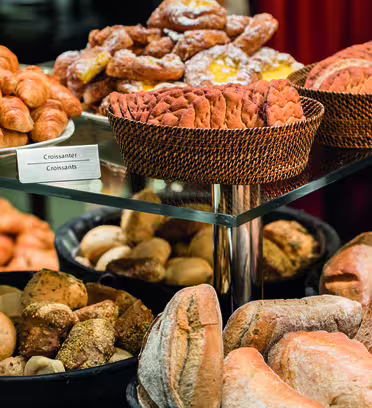 En visning av forskjellige bakevarer, inkludert croissanter, bakverk, rundstykker og brød. Varene er arrangert i kurver og på tallerkener, og viser en rekke teksturer og former i et bakerimiljø.
