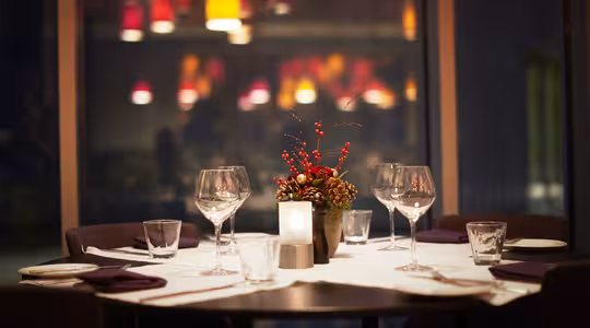Et svakt opplyst restaurantbord for fire med en hvit duk, vinglass og små tallerkener. Et blomstret midtpunkt med røde bær og grønt er i midten. Myke, fargerike lys henger i bakgrunnen.