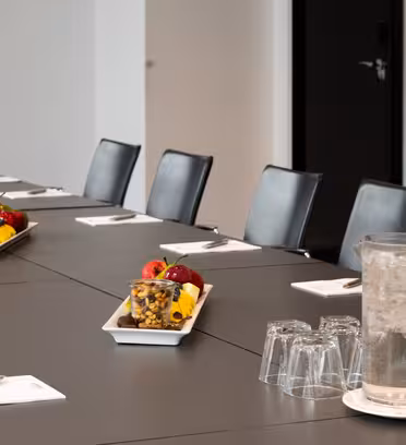 Et konferanserom med et langbord dekket for et møte. Bordet har svarte stoler, notatblokker, penner og brett med frisk frukt og nøtter. En mugge fylt med vann og flere glass er også synlige.