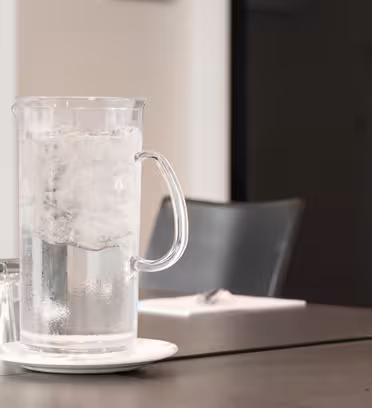 En glassmugge fylt med isvann sitter på en hvit tallerken på et bord. I nærheten er det arrangert flere opp-ned glass. Bakgrunnen viser en uskarp kontorsetting med stoler og en lukket dør.