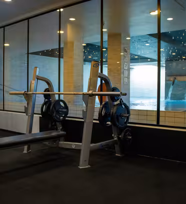 Et treningsområde med benkpress foran store vinduer. Vinduene har utsikt over et innendørsbasseng med brusende vann. Rommet har svart gulv og kremfargede vegger, noe som skaper en moderne og ren atmosfære.