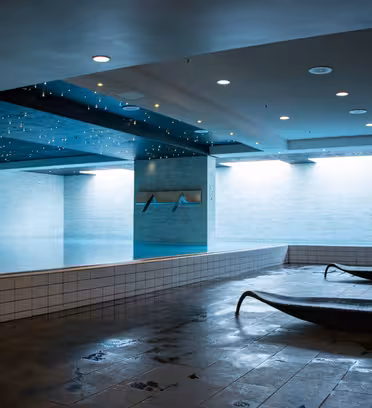 Innendørs bassengområde med moderne design med et rektangulært basseng opplyst av dempet blått lys. Taket er utsmykket med små stjernelignende lys. To mørke lenestoler står på flislagt gulv i nærheten.