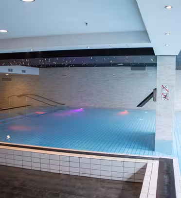 Innendørs svømmebasseng med flislagt gulv, dempet belysning og moderne design. Bassengområdet er omgitt av lyse vegger, med inngangs- og sikkerhetsskilt synlige. Taklysene er på, og bassenget er rolig og ubebodd.