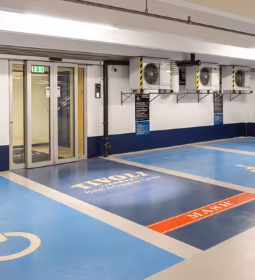 Tilgjengelig parkeringsplass i garasje med to blå rullestol-symbol parkeringsplasser ved siden av en glassdør. Gulvet har "Tivoli" og "Maske" trykket på. Veggmonterte ventilasjonsaggregater er synlige over rommene.