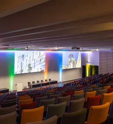 Et tomt, moderne auditorium med rader med flerfargede seter vendt mot en scene. Scenen har flere skjermer som viser arkitektoniske bilder og er opplyst av fargerike LED-lys. Tak og vegger er designet med en elegant, minimalistisk stil.