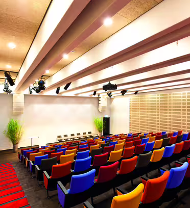 Et moderne, tomt auditorium med rader med fargerike seter vendt mot en scene og en blank skjerm. Taket har trebjelker og det er lys og lydutstyr. Vegger har akustikkplater og det er potteplanter ved scenen.