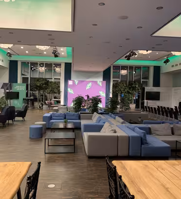 Et romslig arrangementsrom med moderne innredning, med en salong med blå og grå sofaer, trebord og svarte stoler. Grønne planter er spredt utover, med en stor rosa og lilla skjerm foran. Myk grønn belysning lyser opp taket.