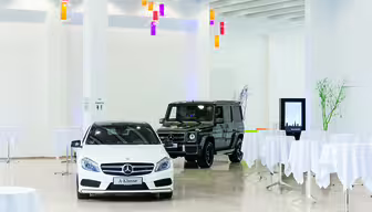 En hvit sedan og en svart SUV vises i et romslig, moderne utstillingsrom med hvite vegger og runde bord dekket med hvite duker. Fargerike pendellamper henger fra taket.