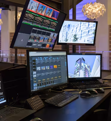 Et kontrollrom med flere skjermer på Tivoli Hotel & Congress Center. Skjermer viser forskjellig innhold, inkludert hotellmerkevarebygging, videoredigeringsprogramvare og arkitektoniske bilder. To hengende dekorative lys er synlige i bakgrunnen.