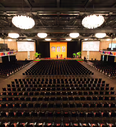 Et stort, tomt auditorium med rader av svarte stoler mot en scene prydet med tre store innrammede bilder. To skjermer flankerer scenen. Lyse lysekroner henger fra taket og lyser opp rommet. Øvre balkonger gir ekstra sitteplasser.
