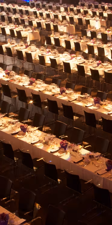 En stor bankettsal er satt opp med mange langbord arrangert i rader. Hvert bord er dekket med hvite duker og dekorert med små lilla floral centerpieces. Svarte stoler står pent på linje langs bordene.